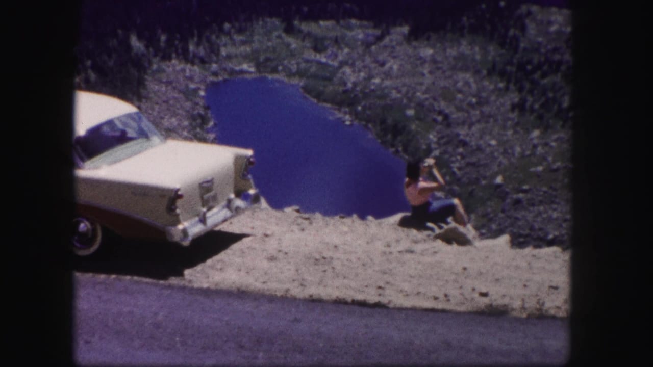 A woman in a red top appears to wave toward the camera from a Colorado lake overlook beside a parked two-tone sedan.