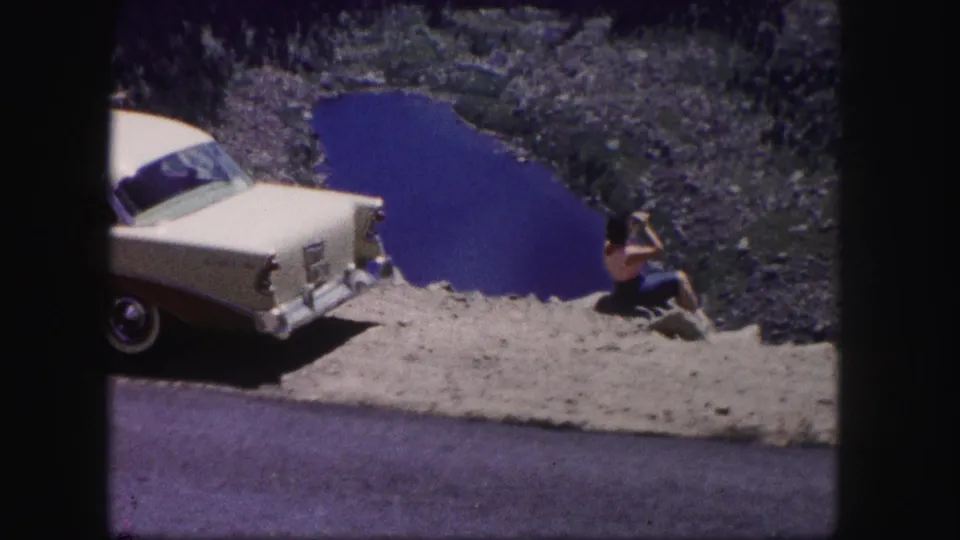 A cream and maroon sedan is parked along a roadside overlook as a woman sits near the ledge above a deep blue Colorado lake.