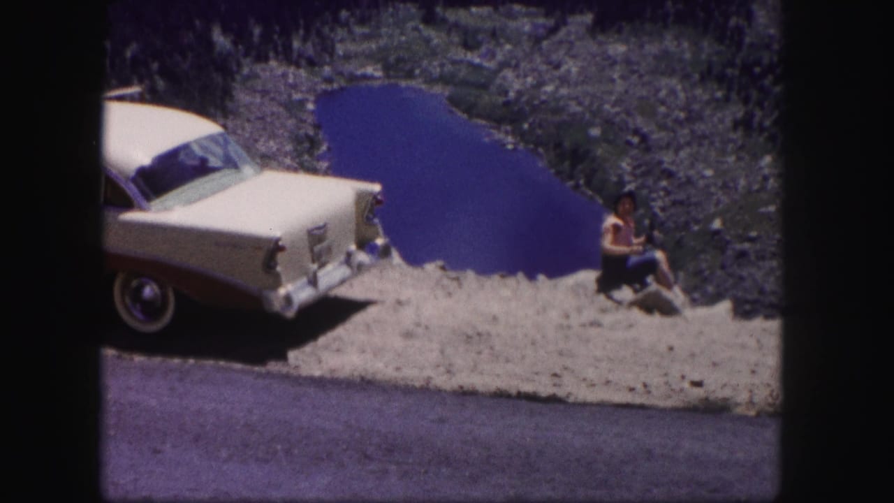 In 1958 Colorado home-movie footage, a woman sits on a rocky ledge beside a cream and maroon sedan overlooking a blue lake.