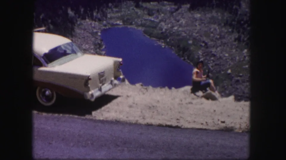 In 1958 Colorado home-movie footage, a woman sits on a rocky ledge beside a cream and maroon sedan overlooking a blue lake.