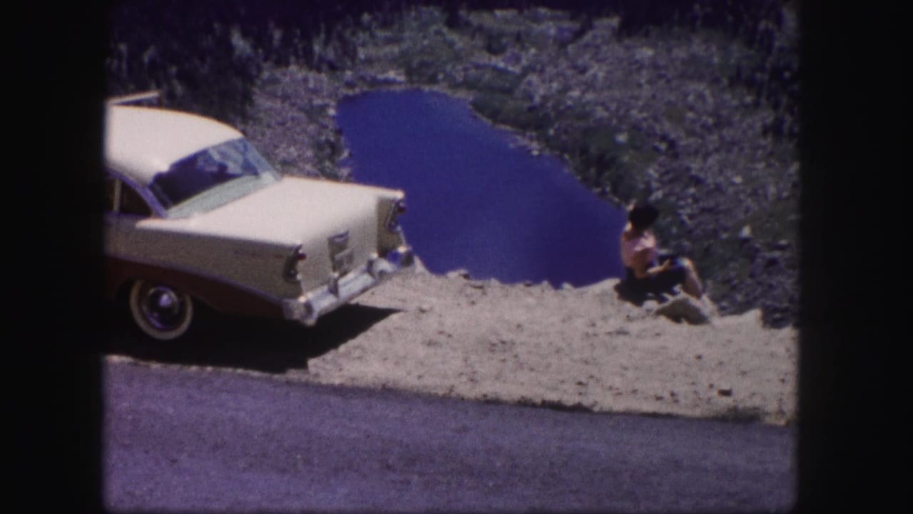 The woman remains seated near the vintage sedan, facing the rocky valley and deep blue lake in the Colorado travel scene.