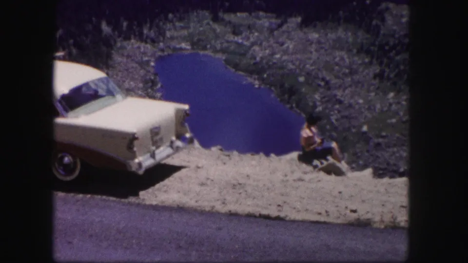A 1950s sedan is parked at the roadside while the woman sits nearby, with the small blue lake and rocky terrain behind her.