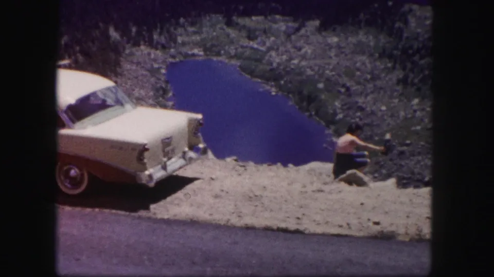 At the end of the clip, the woman shifts or gestures while seated beside the 1950s car overlooking the Colorado lake landscape.