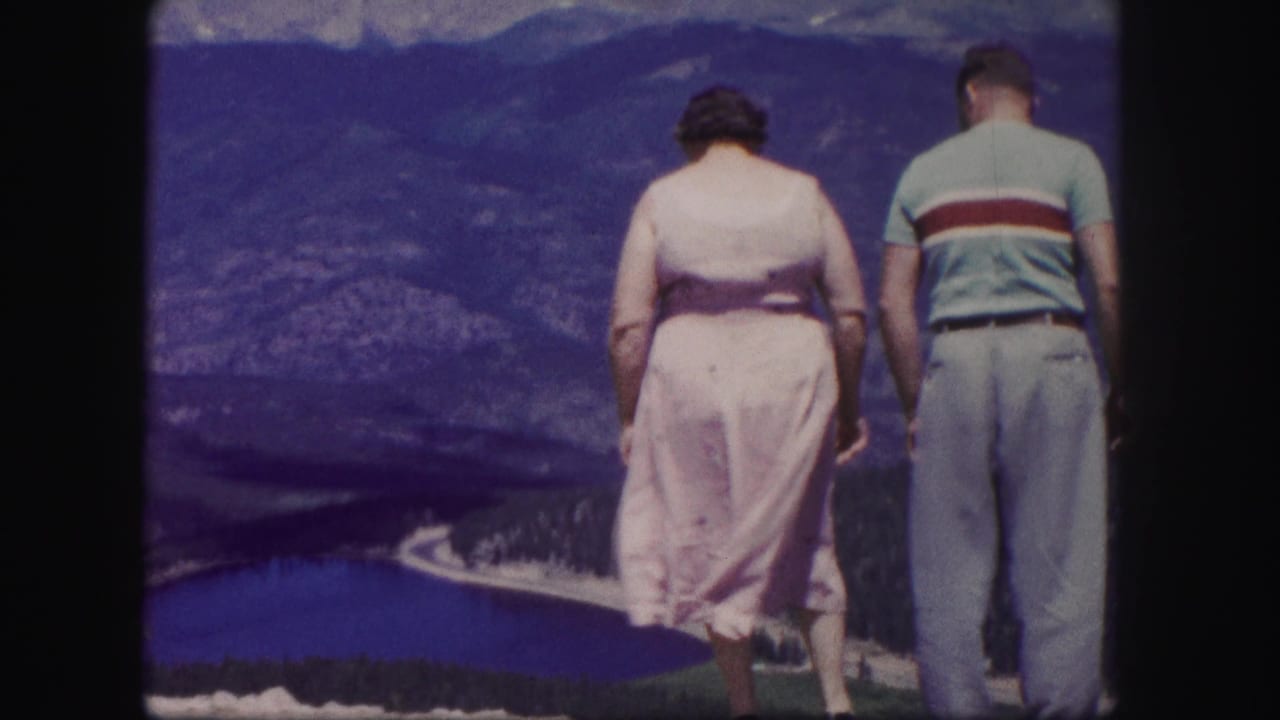 1958 Colorado home-movie frame of a woman in a pale pink dress and a man in a striped shirt looking over a lake and mountains.