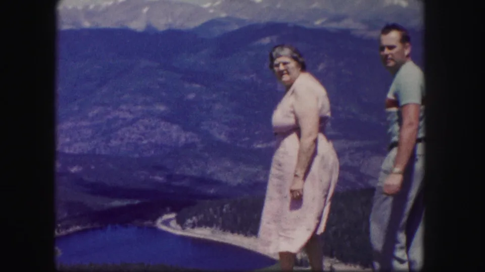 1958 Colorado frame showing the couple turned partly toward the camera at a high overlook above a blue lake and mountain ridges.