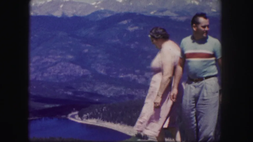 1958 Colorado 8mm frame of the couple moving along the overlook, with a blue lake, wooded slopes, and mountains behind them.