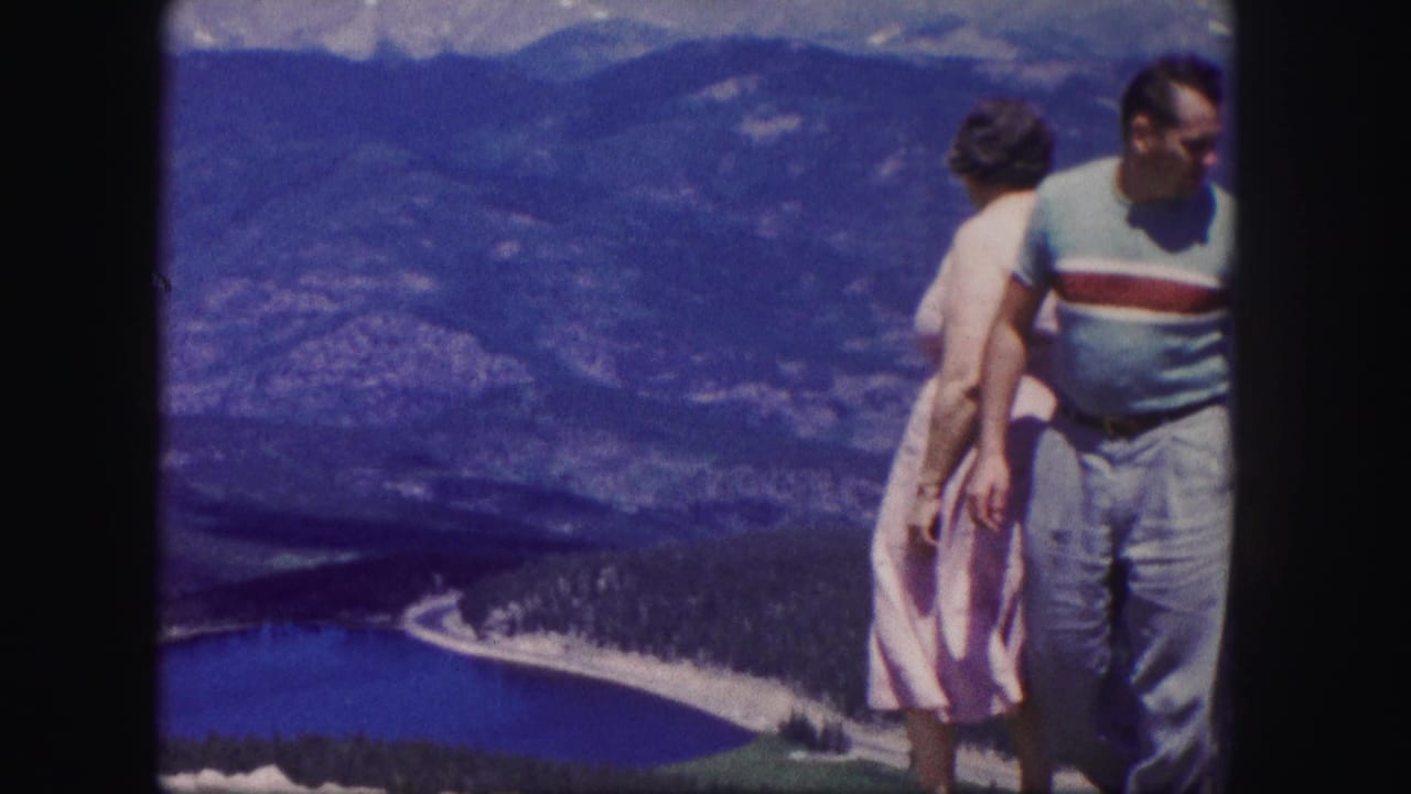 Two adults stand at a mountain overlook above a blue lake and forested valley.