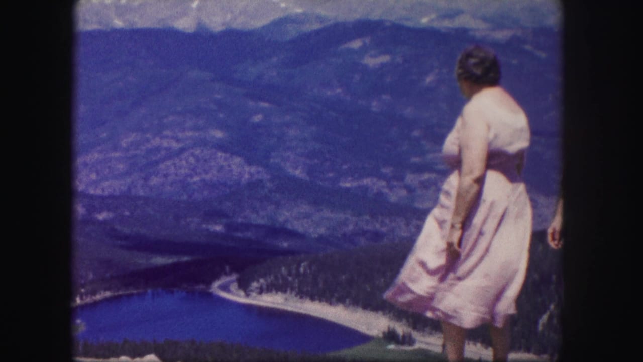 A woman in a light-colored dress stands at a Colorado mountain overlook, looking over a blue lake and distant slopes.
