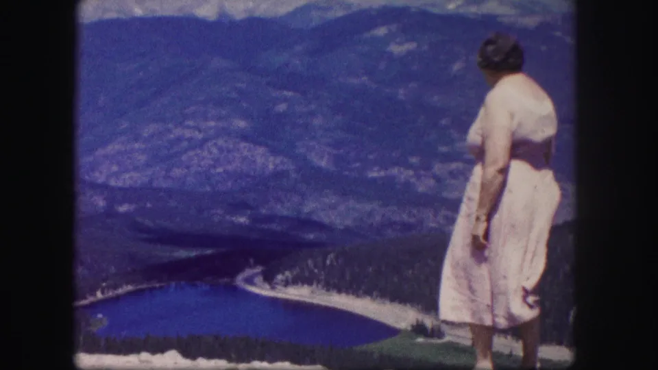 The woman remains at the Colorado overlook, seen in profile against the lake, forested valley, and hazy mountain backdrop.