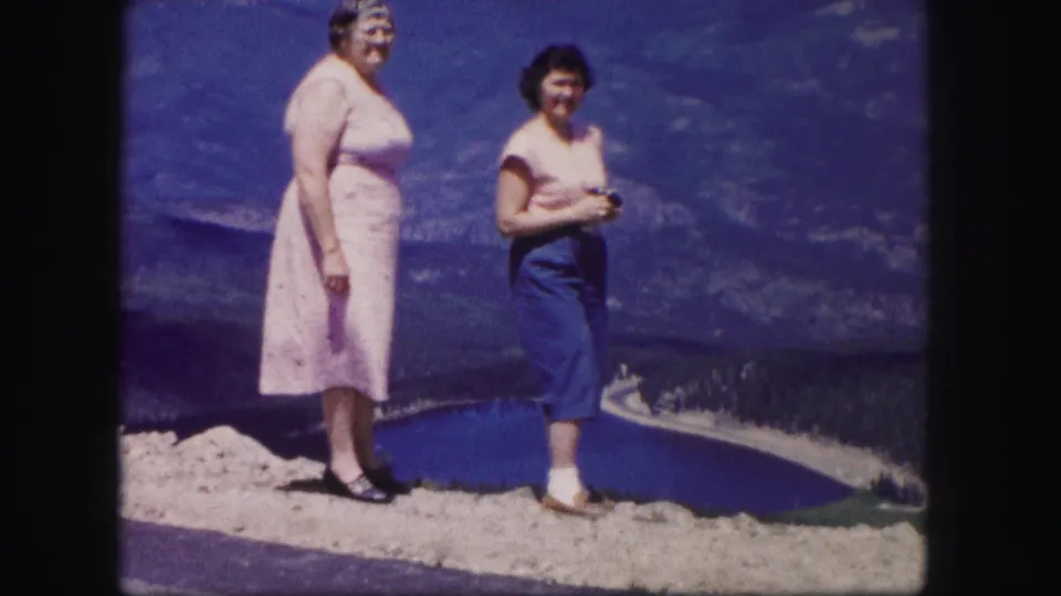 Two women stand at a Colorado scenic overlook in 1958 home-movie footage, with a blue lake and mountain valley in the background.