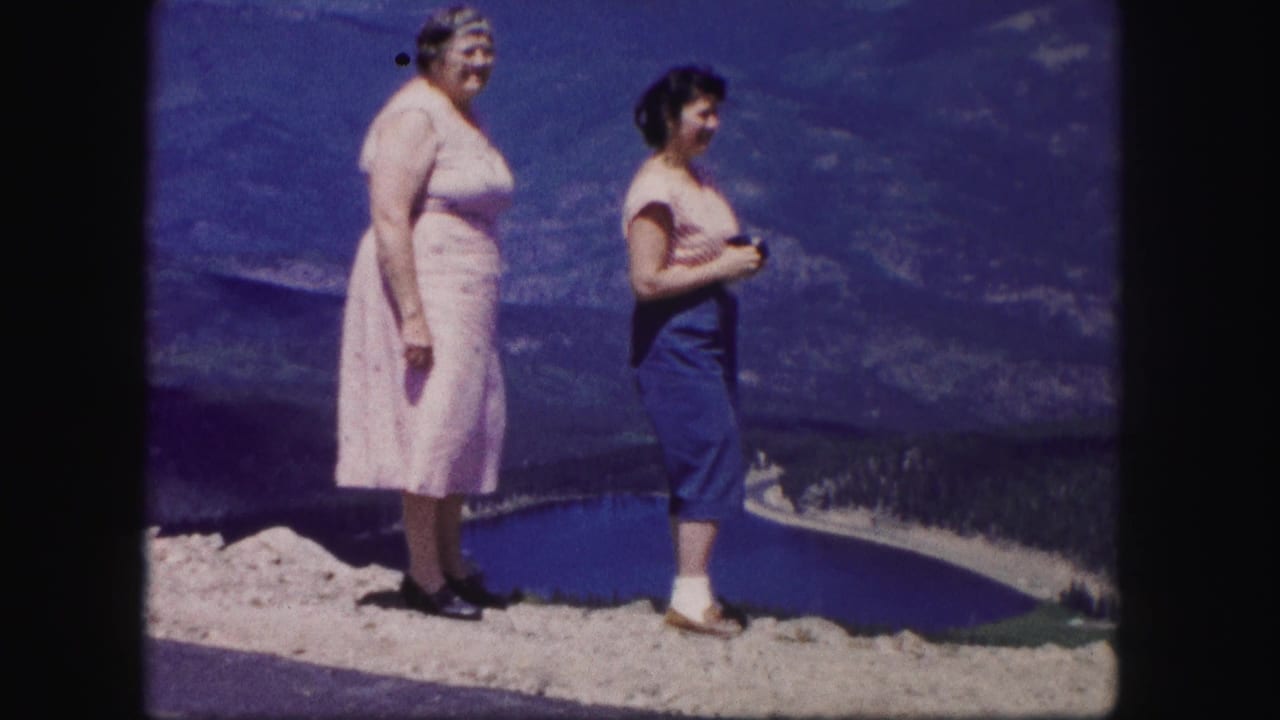 Two women stand on a rocky overlook above a blue mountain lake and distant hills.