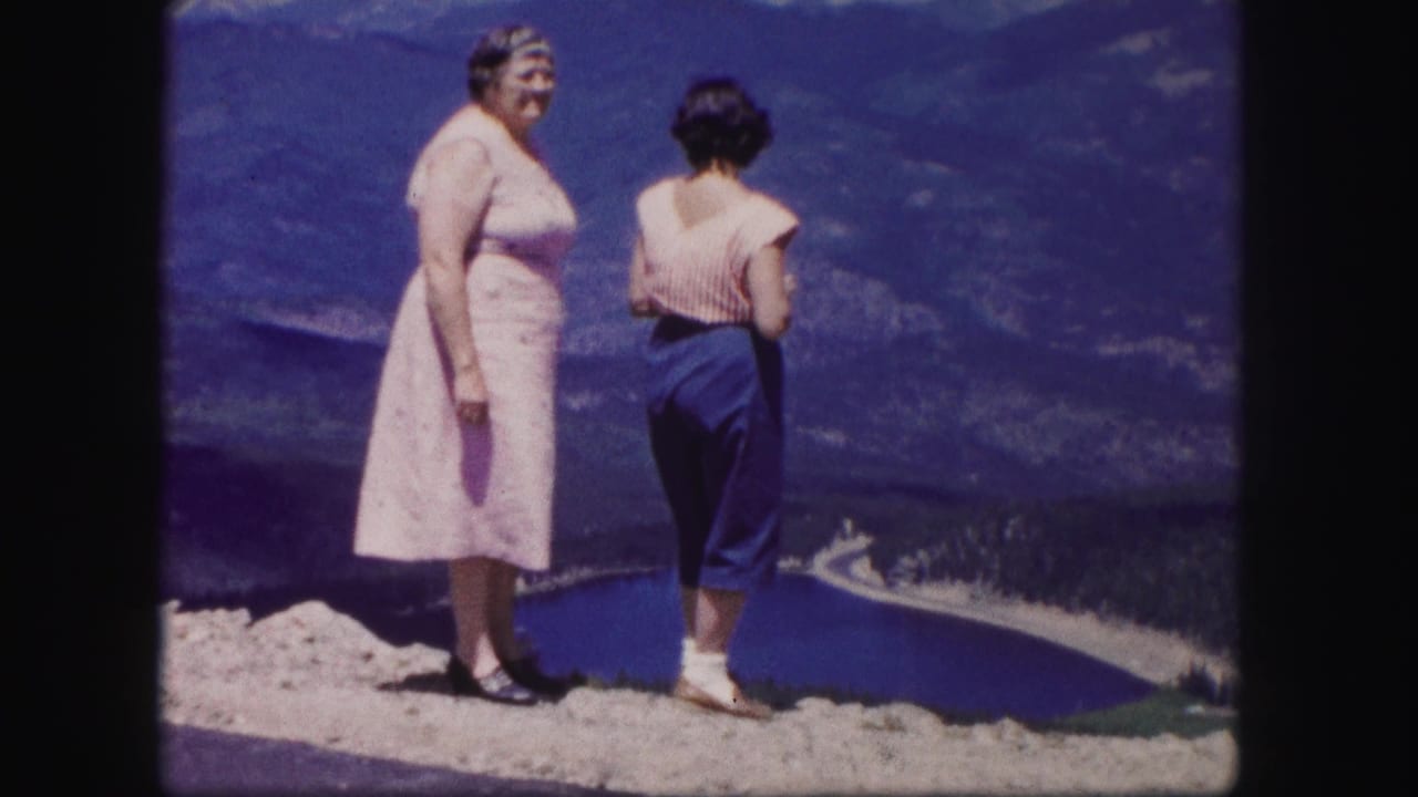 Two women at a Colorado mountain overlook turn toward the lake view, with one woman's back to camera and a curving shore or road below.