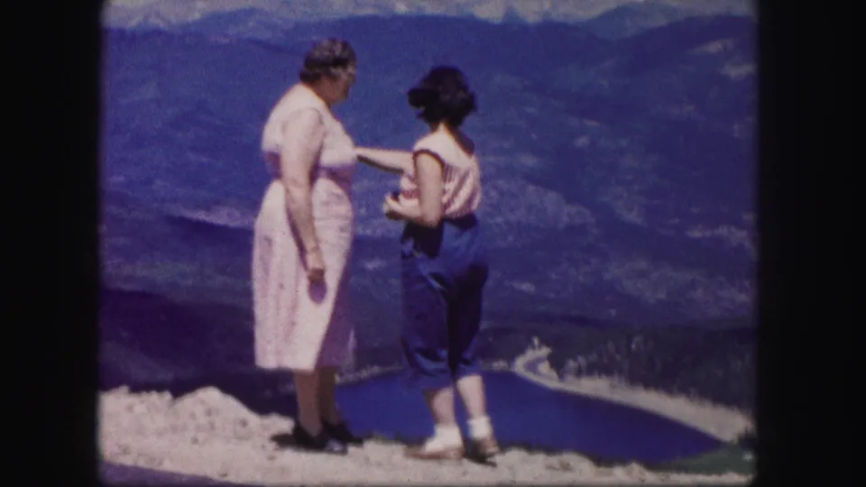 A younger woman gestures toward the Colorado lake and mountain view while an older woman stands beside her on the rocky overlook.
