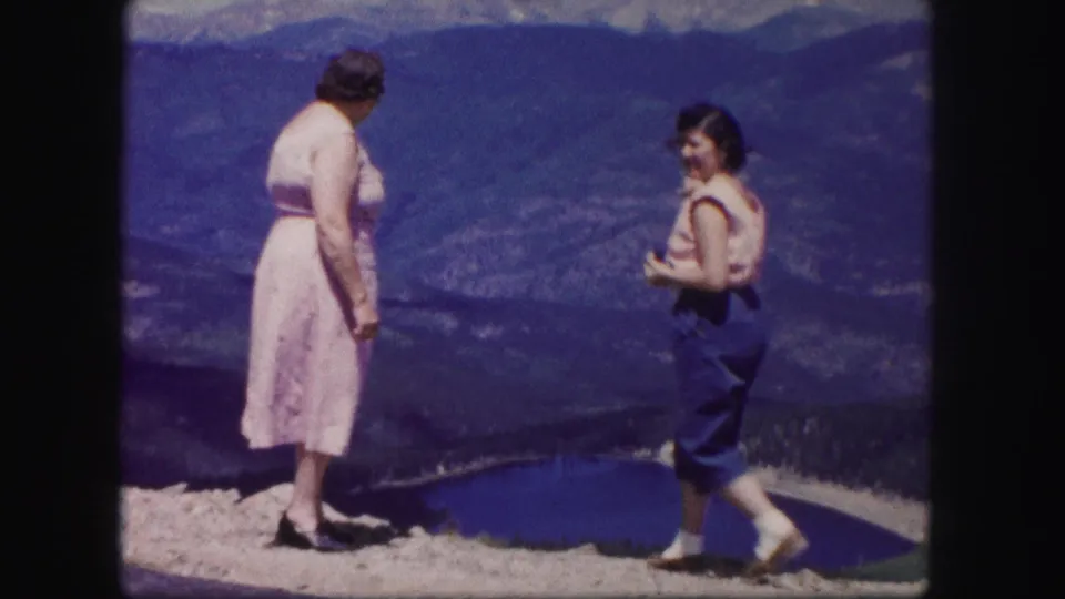 The women stand apart on a Colorado mountain overlook, smiling and turning near a blue lake, forested slopes, and distant ridges.