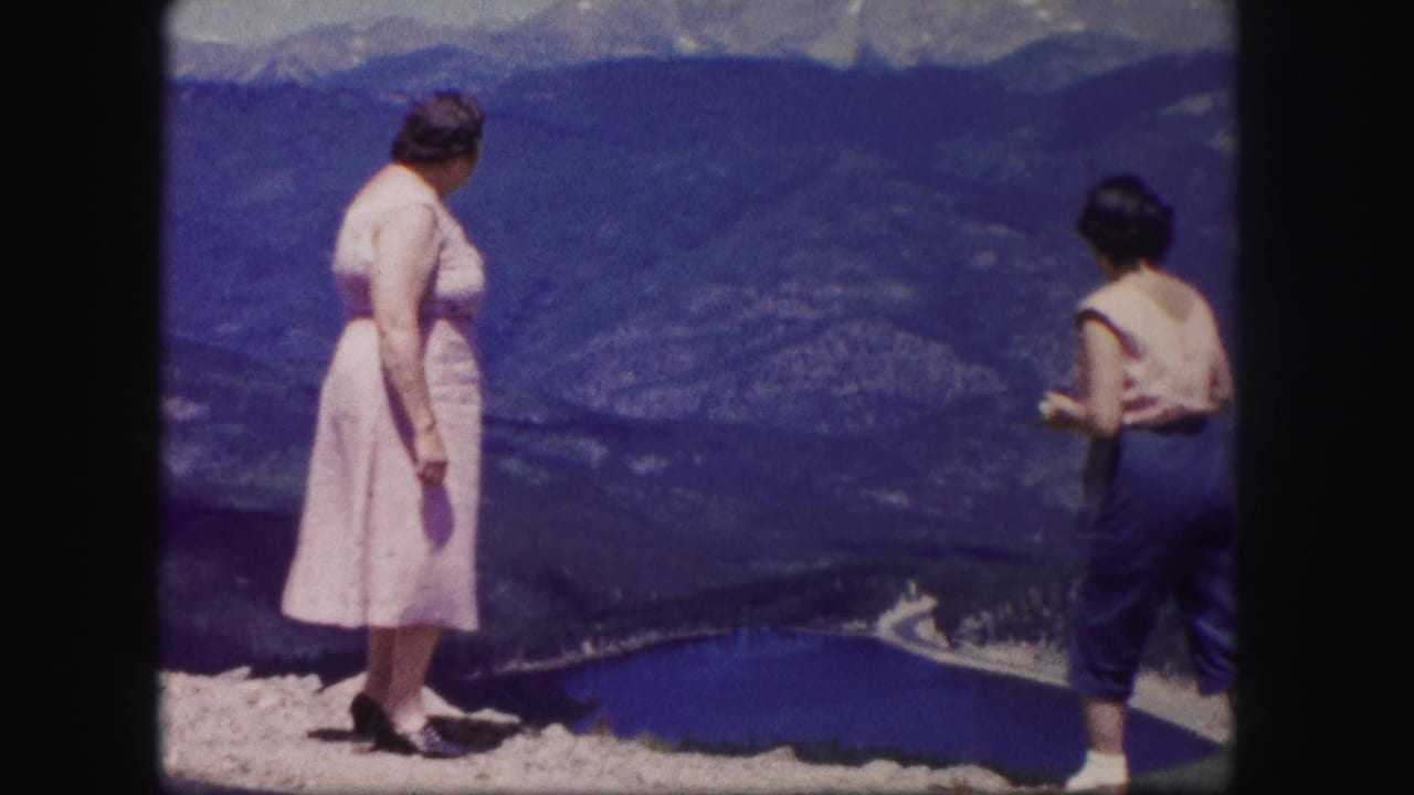 Two women stand on a rocky overlook above a blue lake and mountain valley.