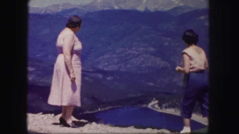 In 1958 Colorado home-movie footage, two women stand at a rocky mountain overlook above a deep blue lake and valley.