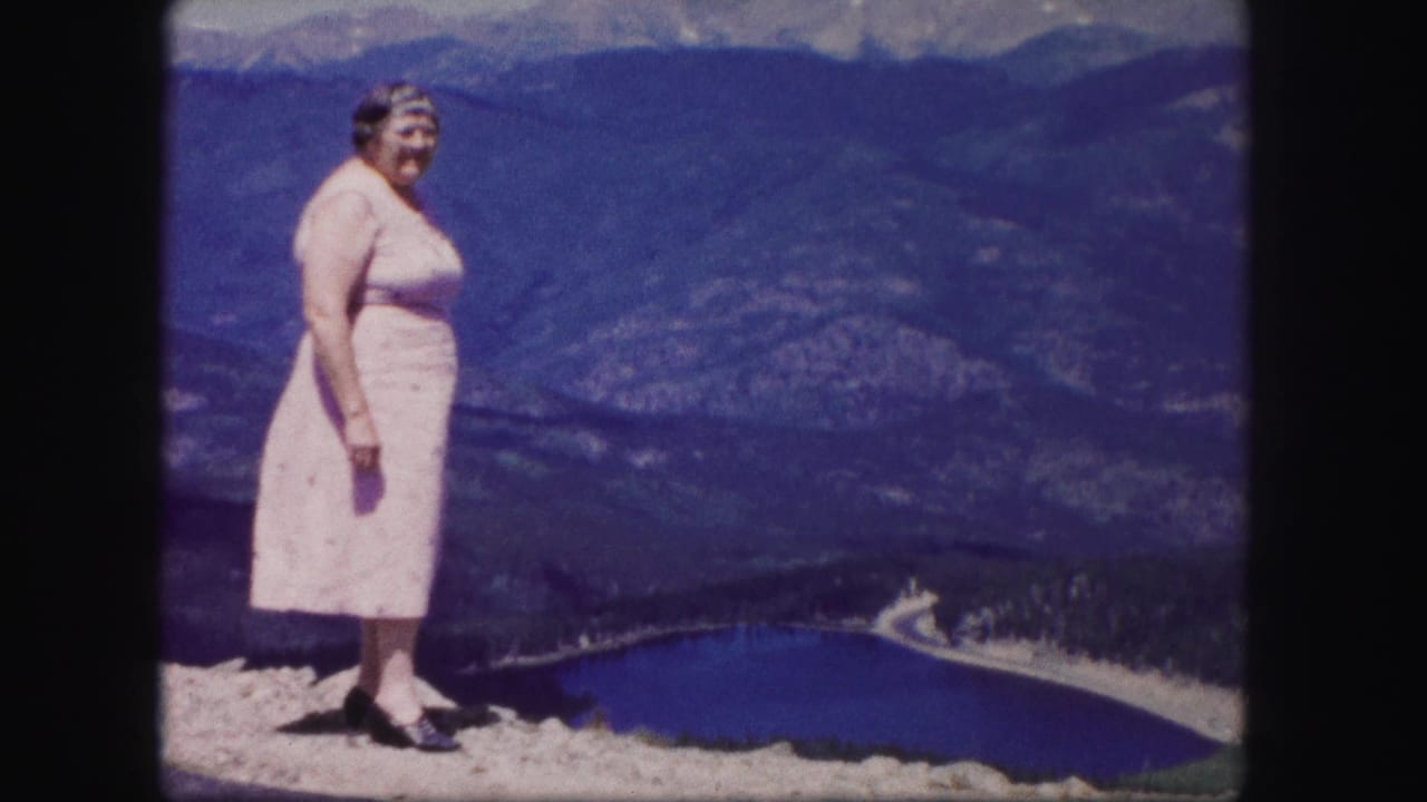A woman in a pink dress stands at a Colorado mountain overlook in 1958, with a lake, valley road, and ridges behind her.