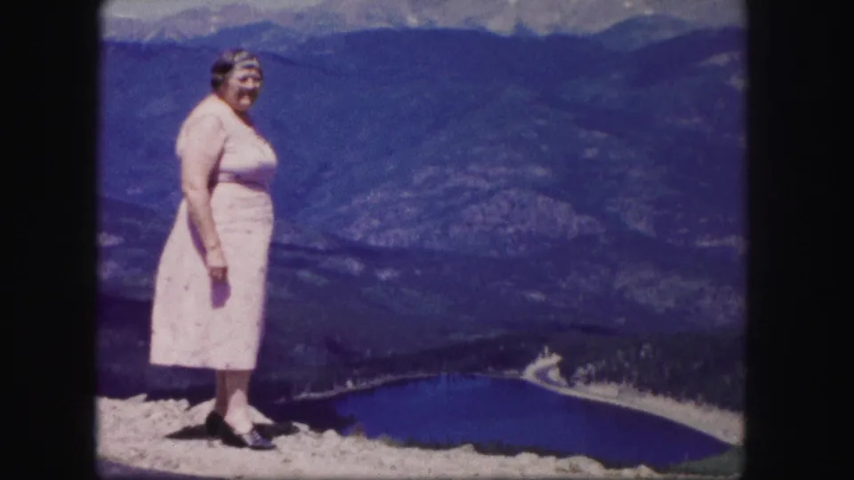 A woman in a pink dress stands at a Colorado mountain overlook in 1958, with a lake, valley road, and ridges behind her.