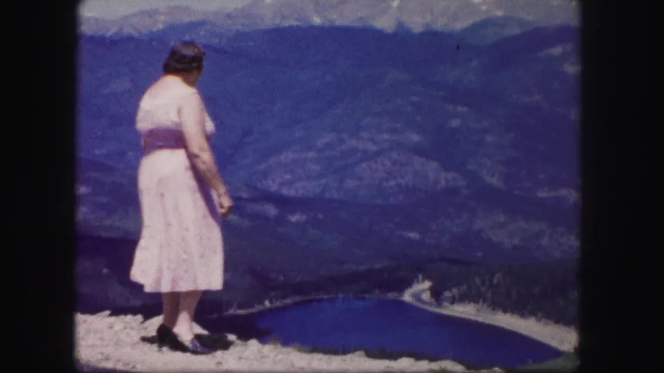 The woman in a pink dress looks toward the blue lake below from a rocky Colorado overlook in 1958 home-movie footage.