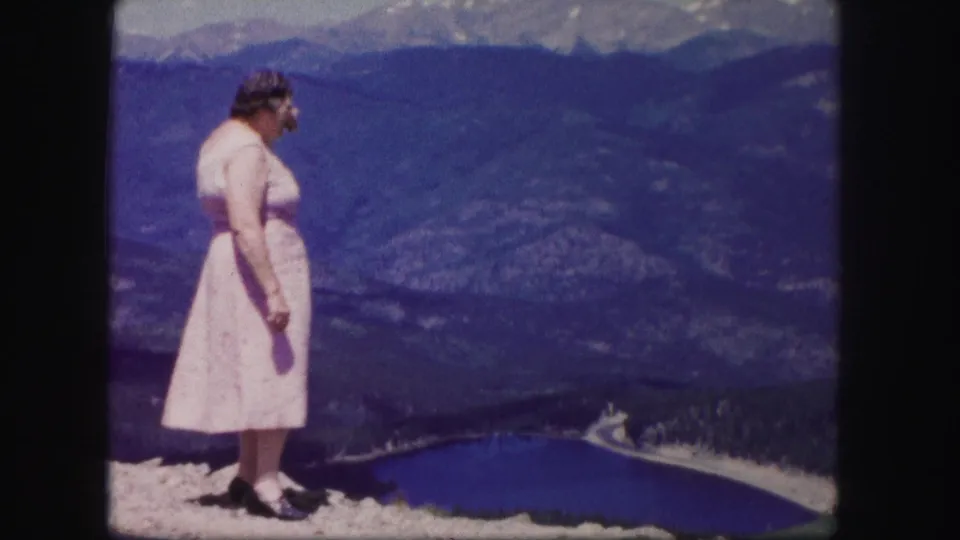 A woman in a pink dress is seen in profile at a 1958 Colorado overlook, with a blue lake and mountain valley behind her.
