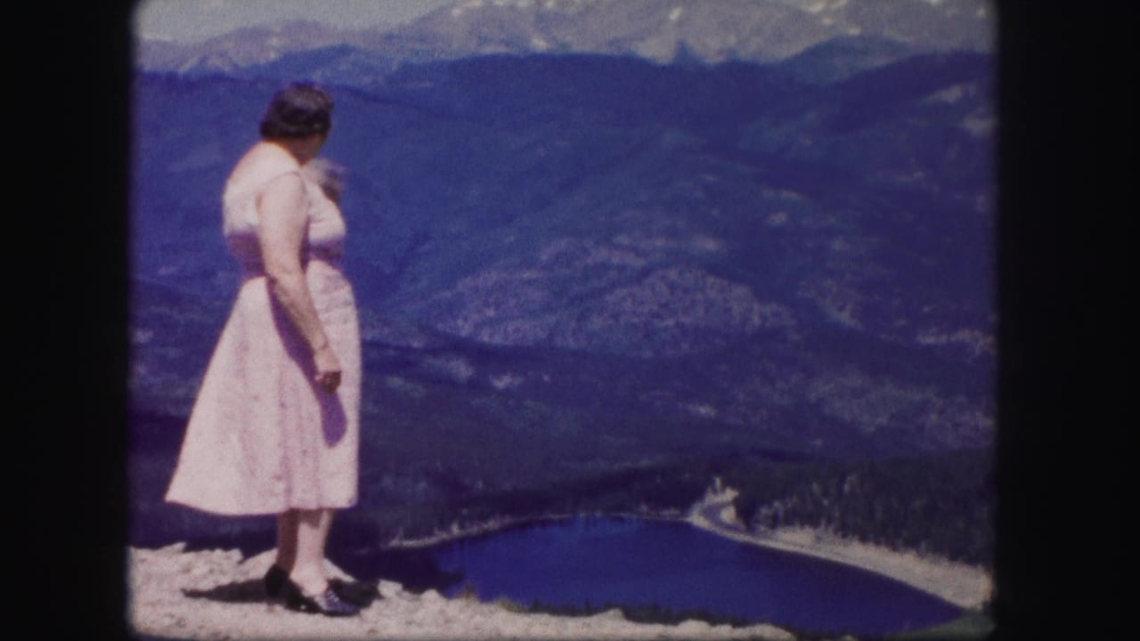 In Colorado 1958 home-movie footage, a woman in a pale dress looks across a blue lake and forested mountain valley from a rocky overlook.