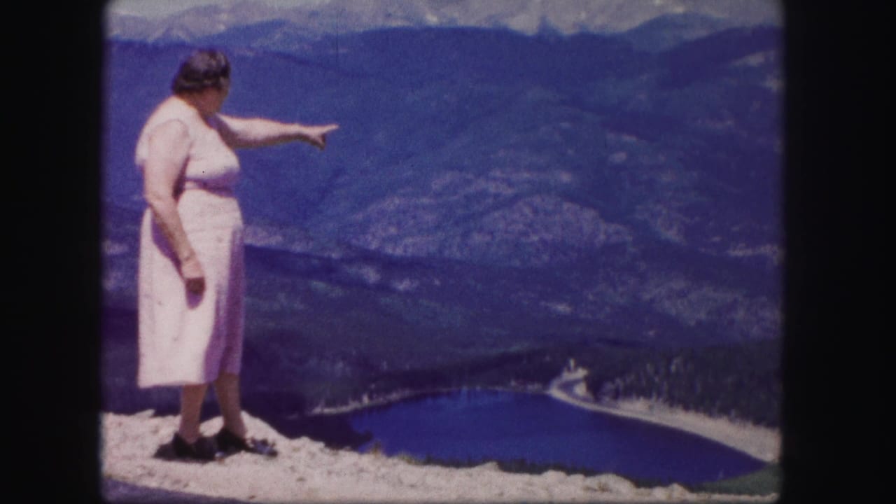 Woman points from a rocky overlook toward the lake and mountain valley below.