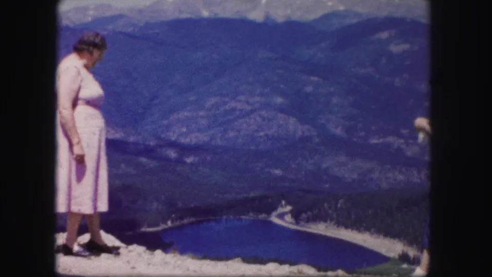 The woman stands near the edge of a Colorado mountain viewpoint, looking down toward a deep blue lake and forested valley in 1958 footage.