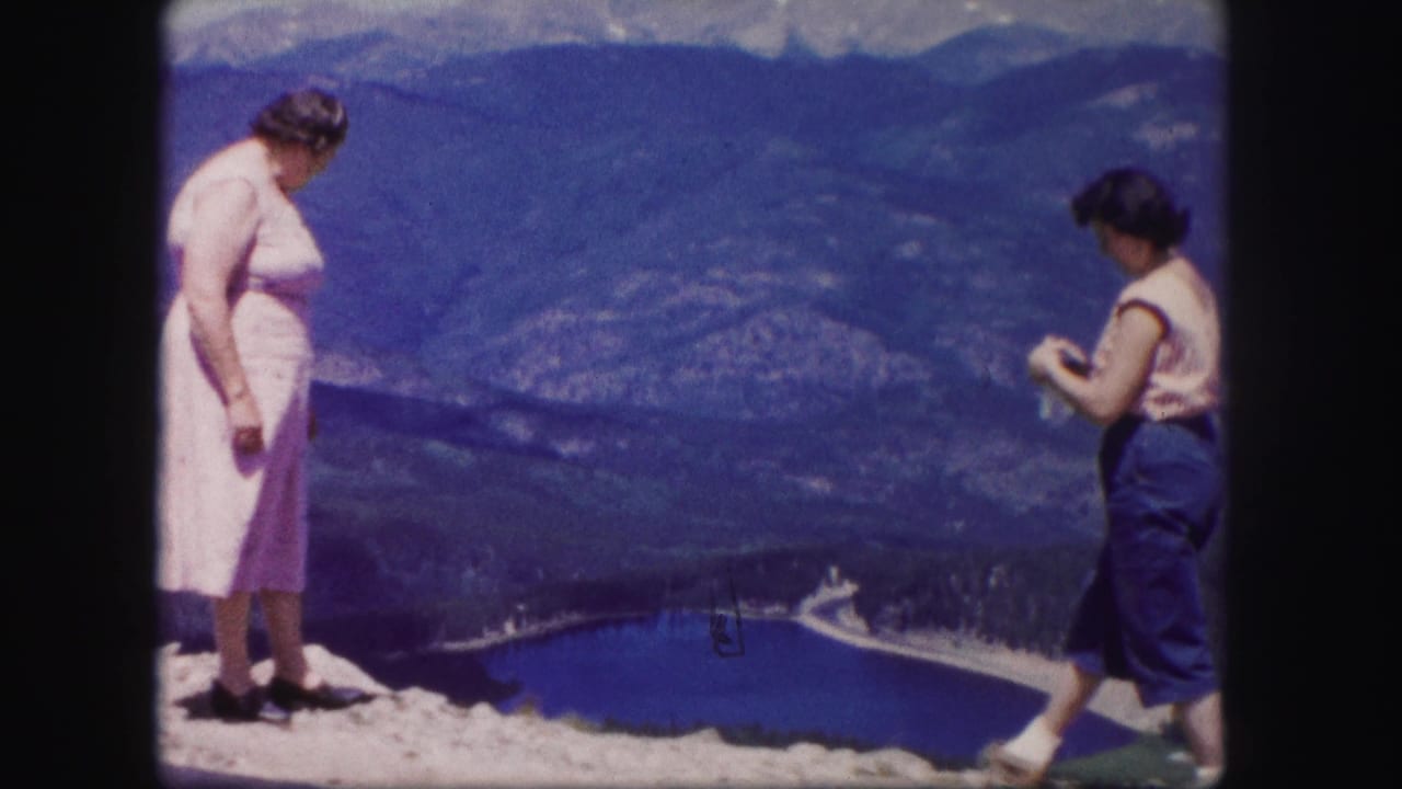 In 1958 Colorado home-movie footage, two women stand at a rocky overlook above a blue lake and distant mountain ridges.