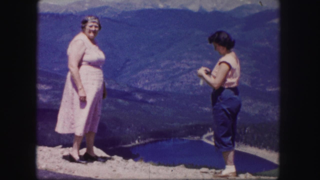 A woman in a pink dress faces the camera while another woman stands near a mountain lake overlook.
