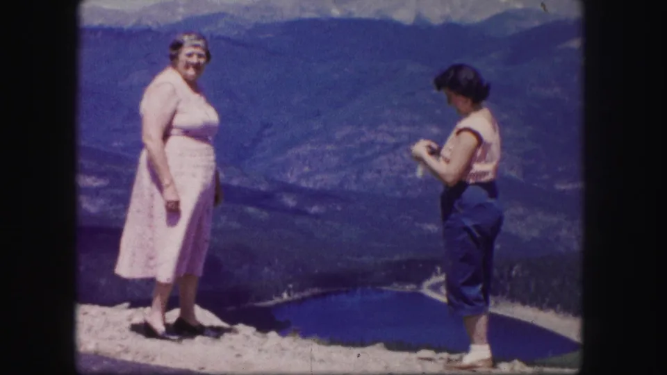 A woman in a pale pink dress faces the camera as another woman stands nearby at a Colorado mountain lake overlook.