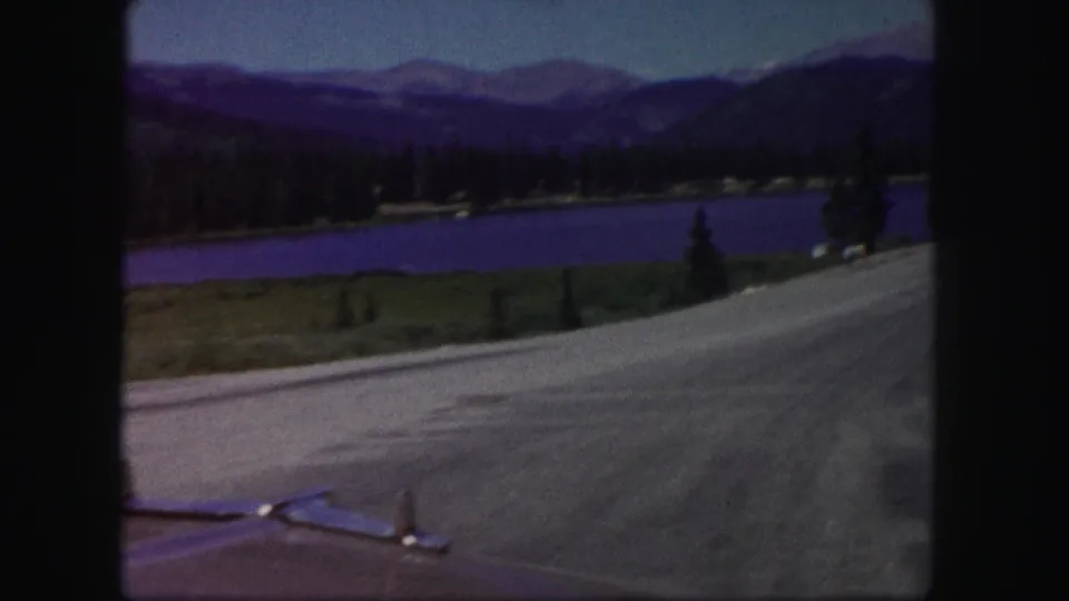 A 1958 Colorado road view shows a car hood in the foreground, a paved road, a blue lake, trees, and mountains beyond.