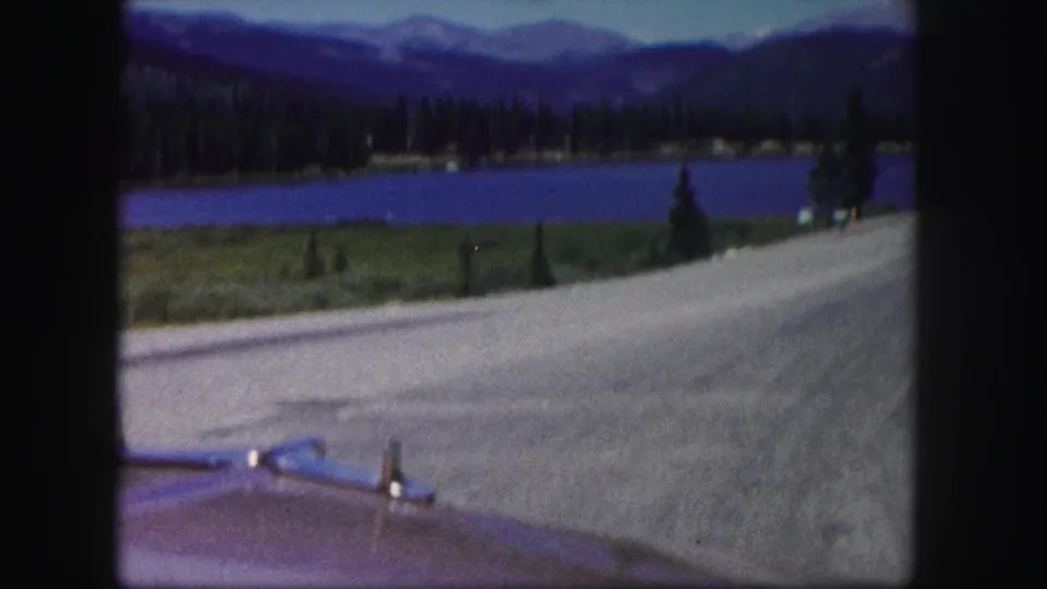 From a moving car, the 1958 Colorado footage looks across a paved road toward a blue lake, forested shore, and mountain backdrop.