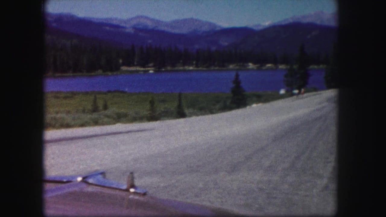 1958 Colorado 8mm frame from a moving car showing a road, lake, evergreen shore, and distant mountains.