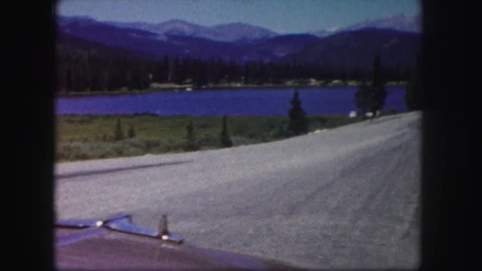 1958 Colorado 8mm frame from a moving car showing a road, lake, evergreen shore, and distant mountains.