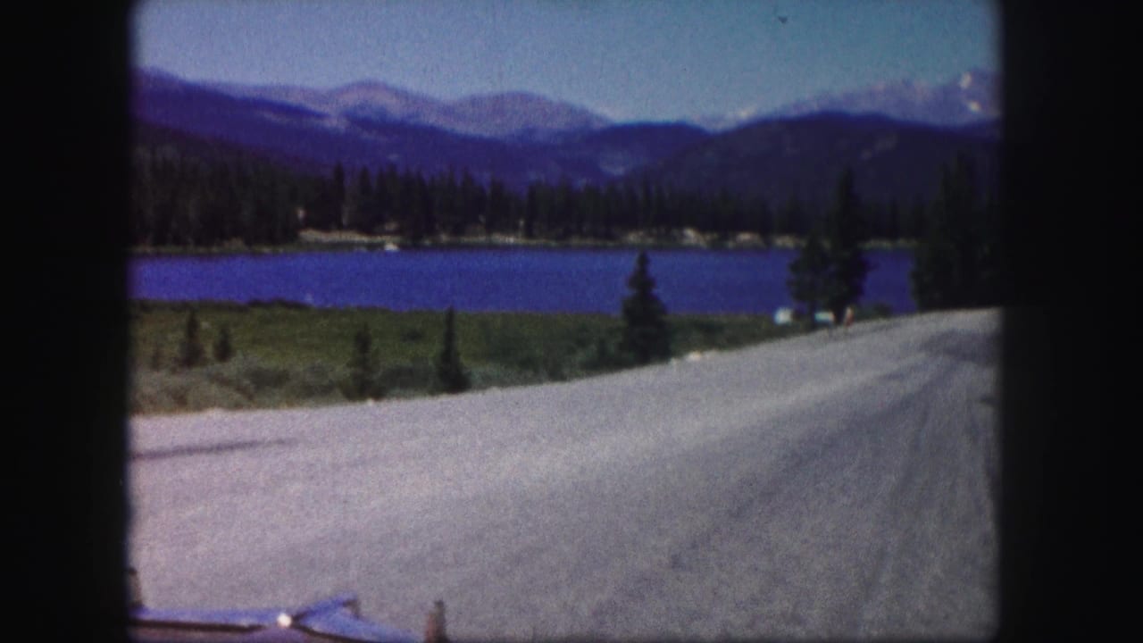 1958 Colorado road-trip frame with a lake, evergreen shoreline, winding road, and distant mountain range.