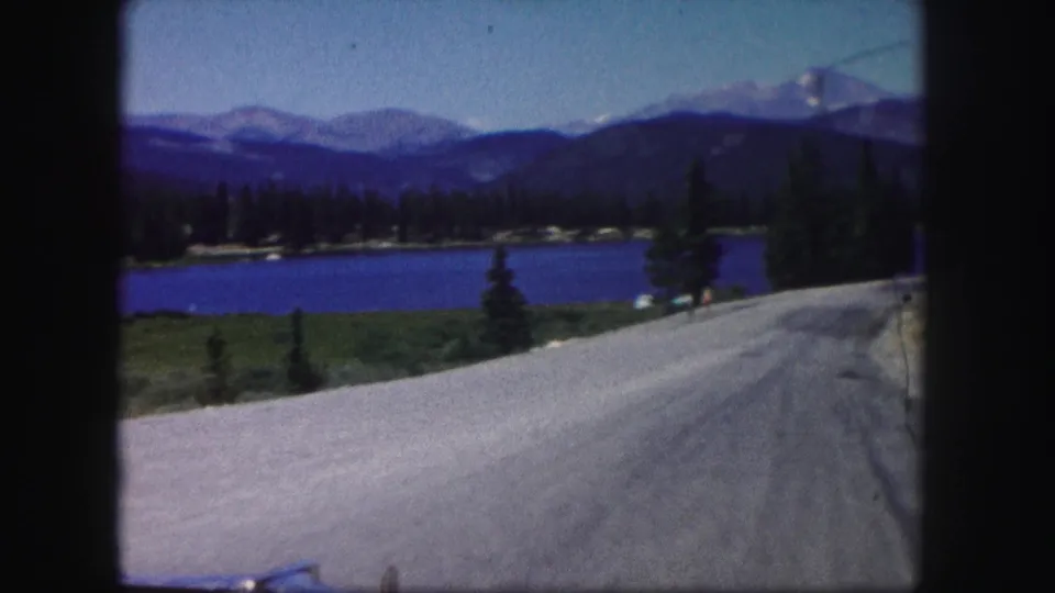 1958 Colorado 8mm view from a moving vehicle showing a road, lake, trees, mountains, and a small distant person.