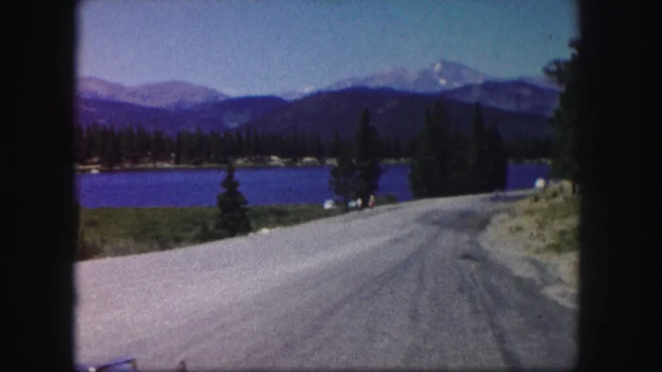 1958 Colorado travel frame showing a road descending past a lake, trees, mountains, and a sloped roadside edge.