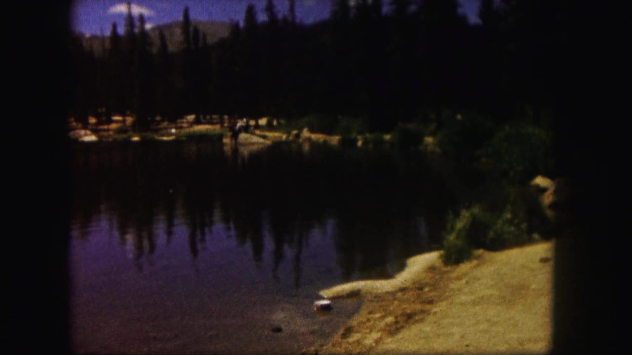 Mountain lake with evergreen trees, reflected shoreline, sandy bank, and dark rounded film frame edges.