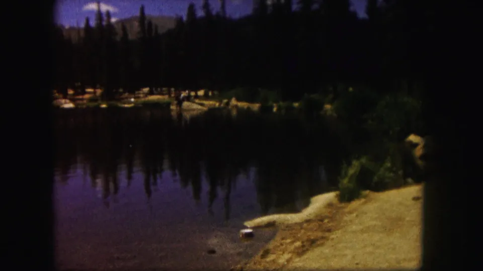 1958 Colorado 8mm frame showing a still mountain lake bordered by evergreens, pale shoreline, rocks, and distant slopes.