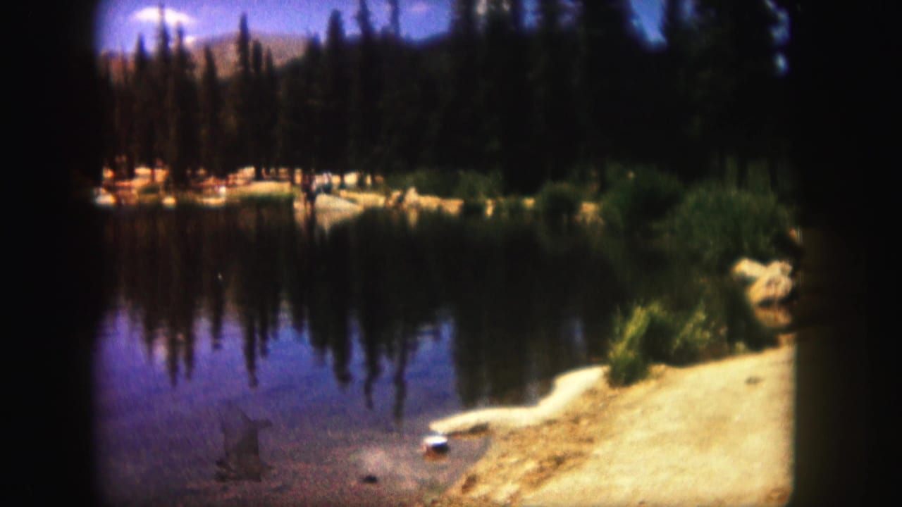 1958 Colorado home-movie view of a calm lake, dark evergreens, sandy shore, rocks, and reflected trees in the water.