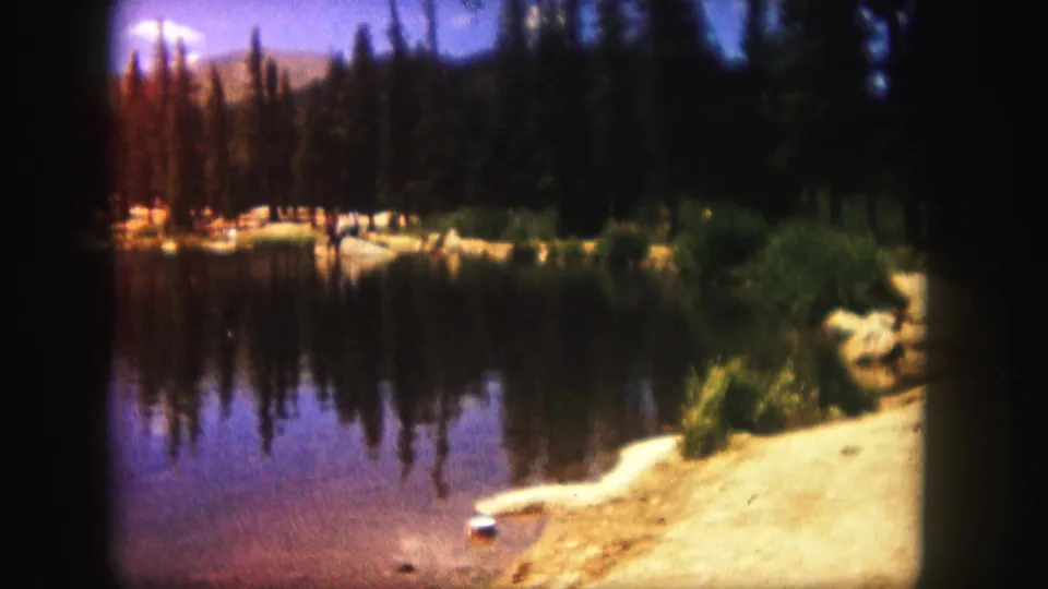 1958 Colorado 8mm footage frames the lake from the shore, with evergreens, reflections, pale sand, and distant terrain.