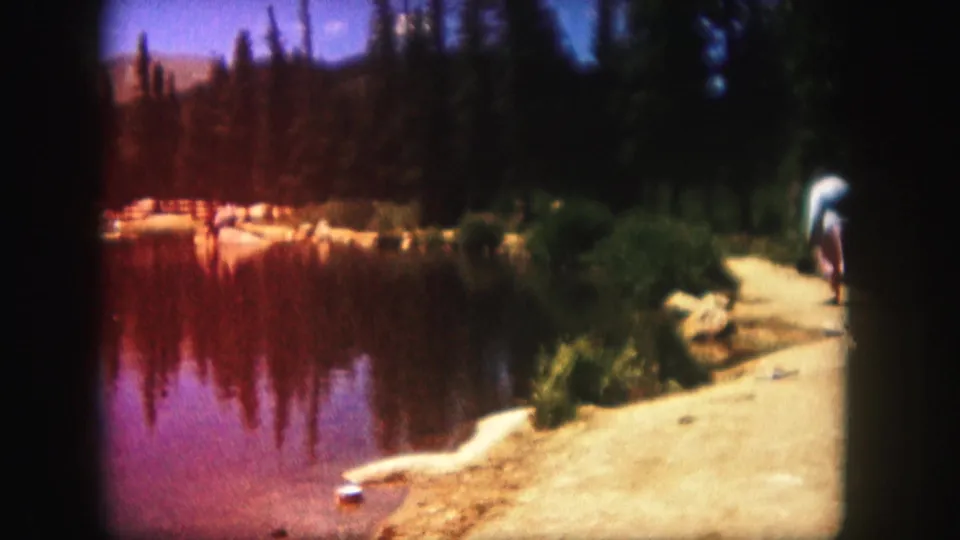 1958 Colorado lake scene with one distant shoreline figure, evergreen trees, sandy bank, rocks, and reddish color shift in the film.