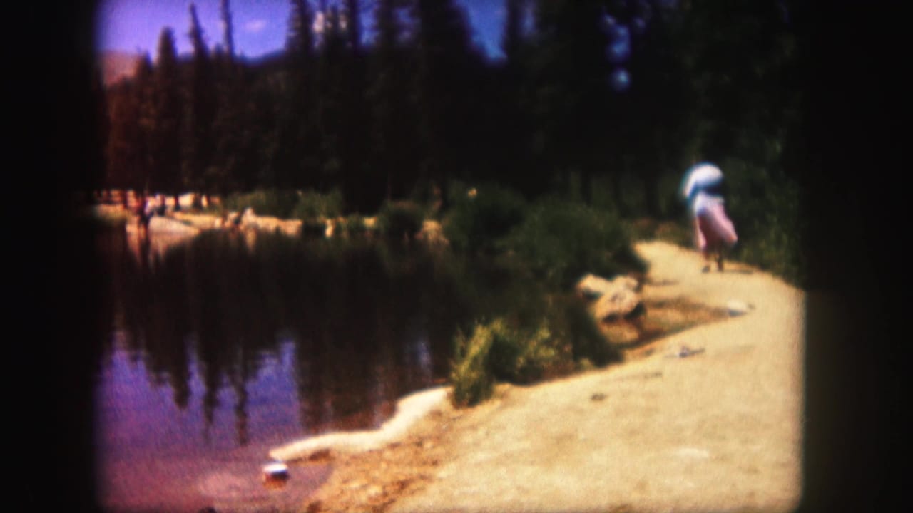 1958 Colorado home-movie frame with a person walking away along a bright lakeside path near still water and dark evergreen trees.