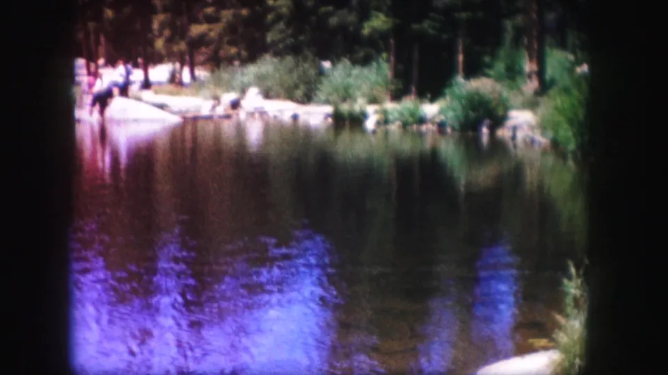 1958 Colorado lakeside frame showing reflective water, shoreline reeds and rocks, evergreen trees, and small distant figures across the lake.