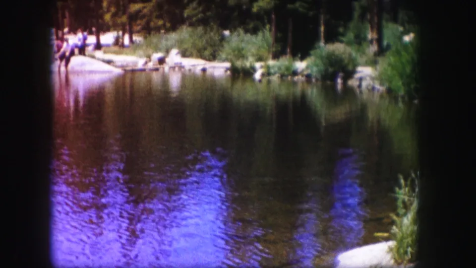 1958 Colorado 8mm frame looking across a reflective lake toward reeds, rocks, evergreen trees, and distant people gathered near the shore.