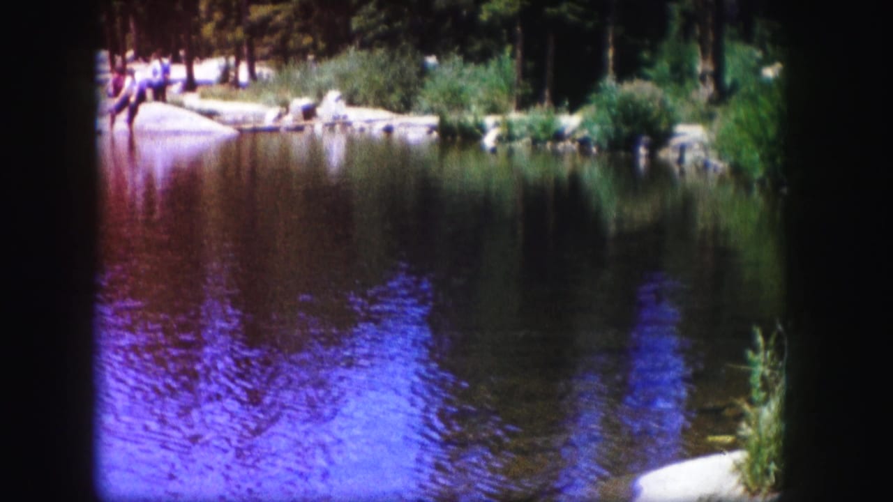 1958 Colorado lakeside view with blue-purple water reflections, reeds, rocks, trees, and small figures near the opposite shore.