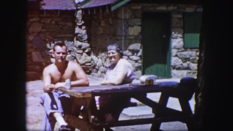 1958 Colorado home-movie frame with a shirtless man and a woman seated at a wooden picnic table outside a rustic stone building.