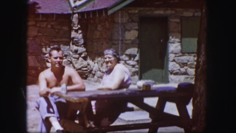 1958 Colorado frame showing two adults seated at a picnic table beside a rustic stone building with a dark doorway and green trim.