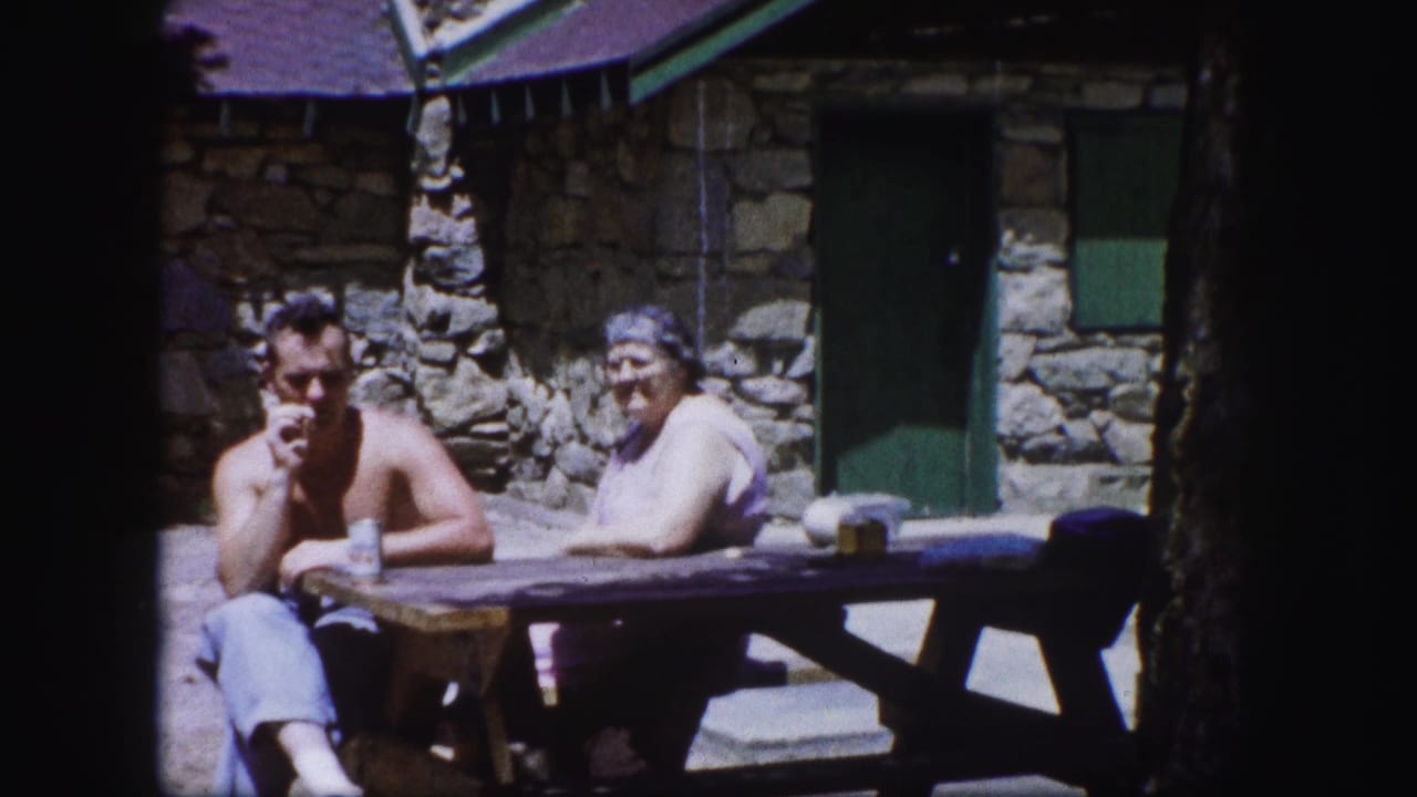 Two adults sit at a wooden picnic table outside a stone cabin.