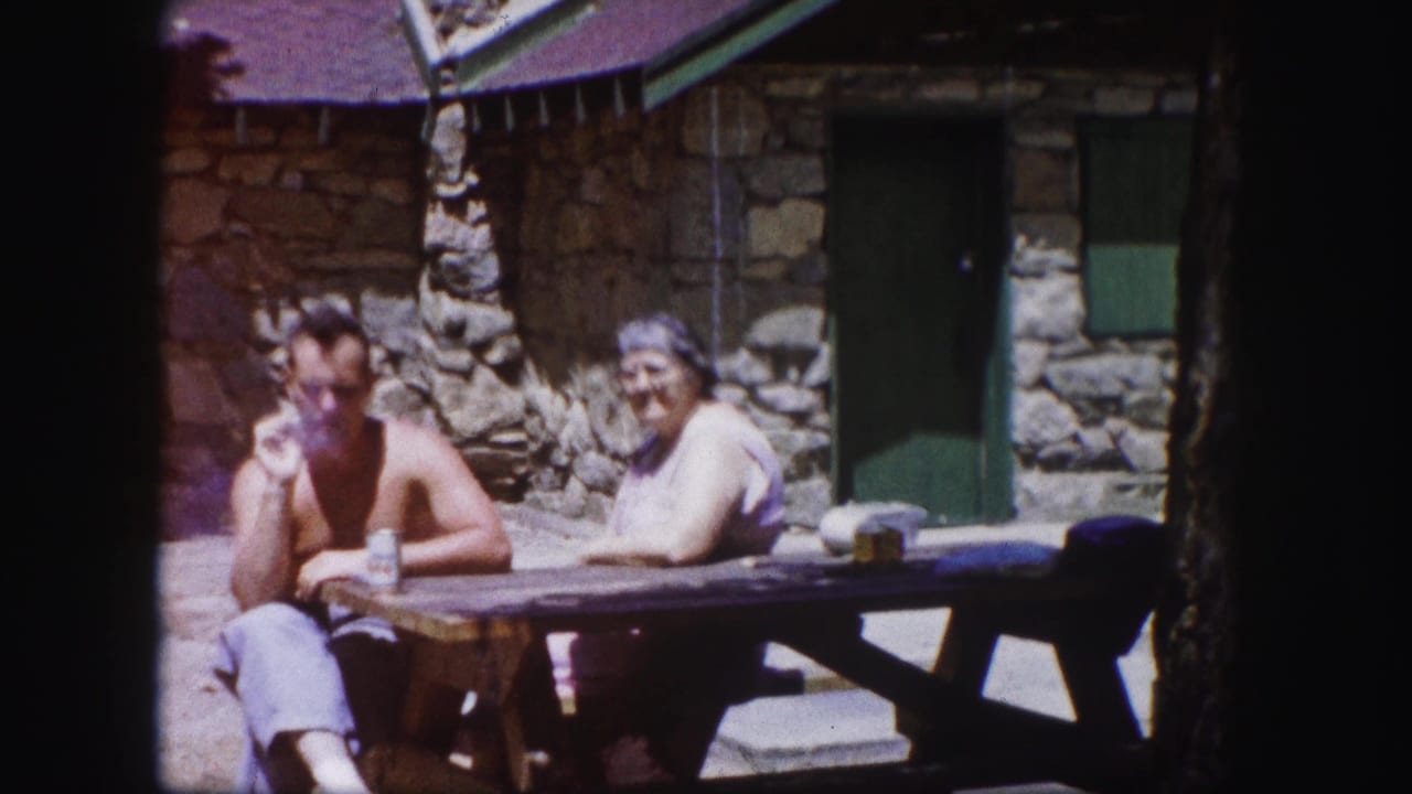 A shirtless man smokes while seated with a woman at a wooden table outside a Colorado stone cabin.